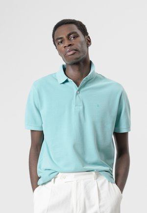 Hombre con polo de manga corta azul claro y pantalones blancos, de pie con las manos en los bolsillos frente a un fondo liso y claro.