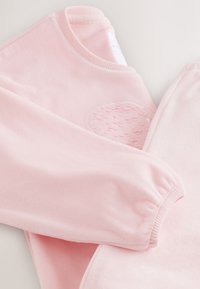 Felpa in pile rosa con una toppa rotonda e testurizzata, polsino elastico e tessuto liscio. Design morbido e leggero.