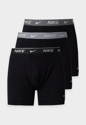 Jeune homme debout pieds nus portant un boxer noir Nike avec une ceinture grise sur un fond blanc uni.