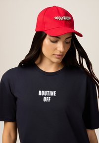 Rode baseballpet met een decoratieve strassaccent aan de voorkant, gedragen met een marineblauwe t-shirt met de tekst "ROUTINE OFF" in het wit.