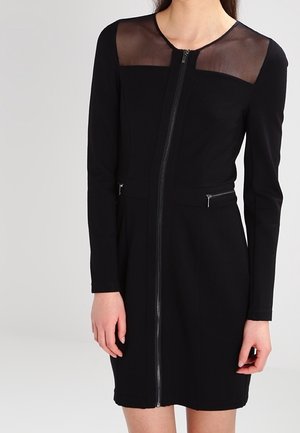 Femme portant une robe noire à manches longues avec une fermeture éclair sur le devant et un panneau transparent sur le haut de la poitrine et les épaules.