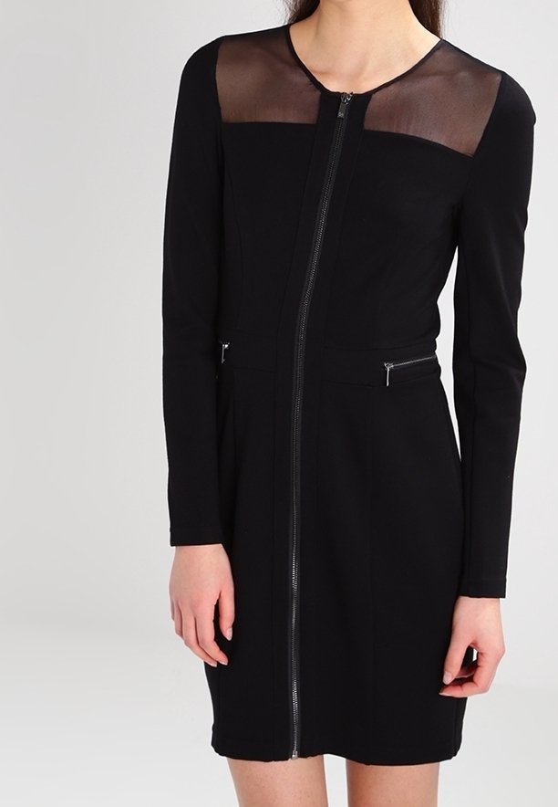 Femme portant une robe noire à manches longues avec une fermeture éclair sur le devant et un panneau transparent sur le haut de la poitrine et les épaules.