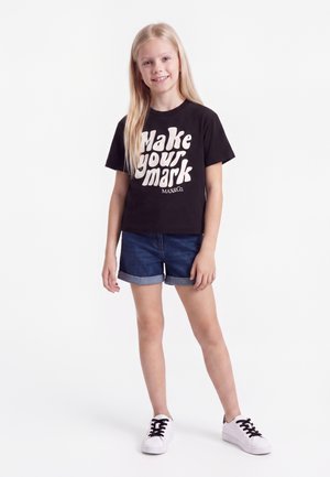 Ung pige med langt blondt hår iført en sort T-shirt med teksten "Make your mark", denim short og hvide sneakers, som står på en hvid baggrund.