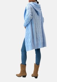 Ljusblå stickad cardigan med huva och kabelstickat mönster, matchad med åtsittande blå jeans och bruna ankelboots, sedd bakifrån.