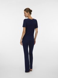 Γυναίκα με τα μαλλιά της σε έναν κότσο, στέκεται ξυπόλυτη, φορώντας έναν συνδυασμό κοντομάνικης μπλούζας και παντελονιού σε σκούρο μπλε χρώμα, σε ένα απλό λευκό φόντο.