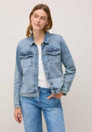 Eine Frau in einer hellen Jeansjacke und einem weißen Shirt steht mit der Hand in ihrer Tasche und schaut leicht zur Seite vor einem neutralen Hintergrund.