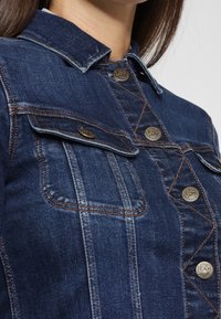 Chaqueta de denim en azul oscuro con un collar clásico, botones metálicos, dos bolsillos en el pecho y detalles de costura en contraste.