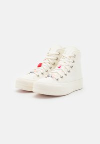 Zapatillas blancas de caña alta con suela de goma texturizada, que presentan acentos de tela de colores y un detalle de letras hecho a mano en la punta.