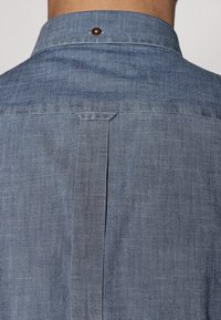 Camisa de mezclilla vista desde atrás, color azul claro, con un botón en el cuello, canesú rectangular y tela texturizada.