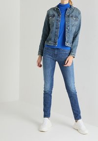 Giacca di denim di un blu medio, con due tasche sul petto e bottoni metallici, abbinata a un dolcevita blu e jeans skinny, completata da sneakers bianche.
