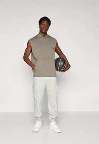 Jovem em hoodie sem mangas e calças de treino, segurando uma bola de basquete preta, de pé contra um fundo claro e liso.