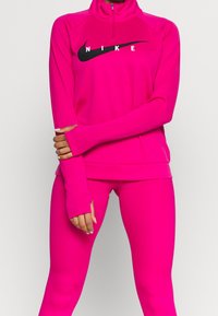 Fuchsia Zip-Hoodie und passende Leggings aus dehnbarem Material mit einem schwarzen Nike-Logo über der Brust und Daumenlöchern in den Ärmeln.