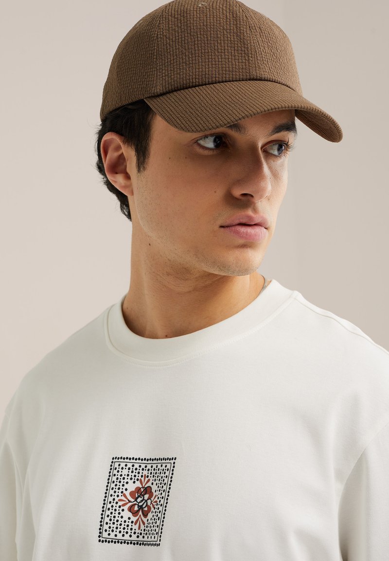 Jeune homme regardant vers la droite, portant une casquette texturée marron et un sweat-shirt blanc avec un petit motif floral abstrait sur la poitrine.