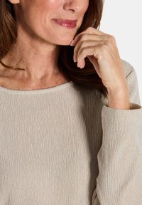 Nahaufnahme einer Frau, die sanft lächelt, während sie ihre Hand an ihr Kinn hält, gekleidet in einem strukturierten, beigen Langarmoberteil.
