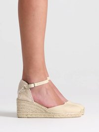 Frau trägt beigen Keil-Espadrille mit Knöchelriemen und geflochtenem Absatzdetail, vor hellem, schlichtem Hintergrund.