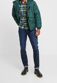 Fyllig grön jacka med huva, blå och gul rutad skjorta, mörka jeans och svarta ankelboots. Jackan har dragkedja och kardborreknäppning.