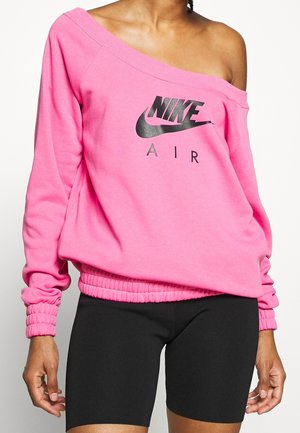 Osoba nosząca jaskraworóżową bluzę Nike Air z odkrytymi ramionami, w połączeniu z czarnymi dopasowanymi spodenkami rowerowymi, stojąca na jednolitym tle.