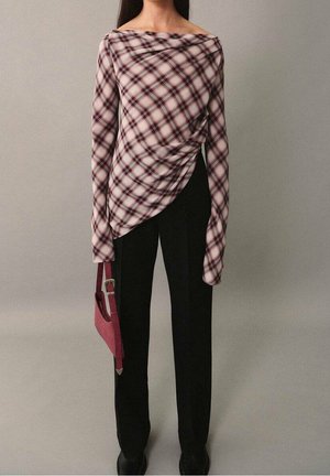 Kobieta w asymetrycznej bluzce w różowo-czarną kratę z długim rękawem, czarnych spodniach, czarnych butach, trzymająca małą różową torebkę.