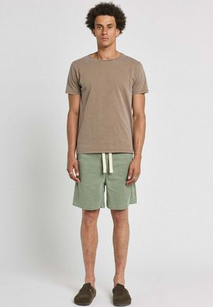 Homme aux cheveux bouclés debout, portant une chemise marron à manches courtes, un short vert à cordon et des chaussures marron sans lacets sur un fond uni.