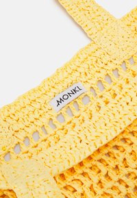 Sac fourre-tout jaune tissé avec une tresse ouverte, doté de larges bretelles et d'une étiquette blanche avec le logo "MONKI". Matériau texturé et durable.
