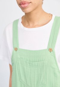 Lichtgroene katoenen tuinbroek met een gestructureerde afwerking, voorzien van houten knopen en een voorzak, gedragen over een wit T-shirt.