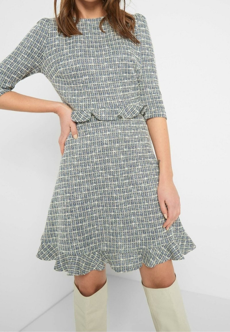 Sininen ja valkoinen ruudullinen mekko, jossa on kolmivartiset hihat, liehuva helma ja röyhelödetaljilla varustettu vyötärö, valmistettu teksturoidusta kankaasta.