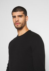 Suéter negro de punto de manga larga con cuello redondo y textura suave, mostrado en un modelo que mira ligeramente hacia la derecha sobre un fondo claro.