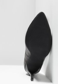 Chaussure à talon haut noire avec un motif texturé en peau de serpent, présentée semelle vers le haut sur un fond blanc en coin.