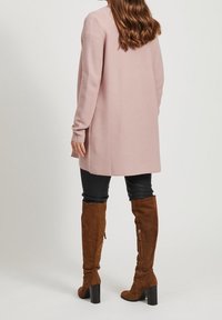 Světle růžový žebrovaný kardigan s dlouhými rukávy a rovný střih, zkombinovaný s černými koženými kalhotami a hnědými semišovými botami do kolen.