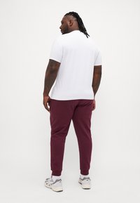 Hvit kortermet skjorte, burgunder joggebukser med mansjetter, grå sneakers, synlige tatoveringer, står med ryggen til kameraet mot en hvit bakgrunn.