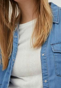 Chemise en jean boutonnée bleu clair, portée sur un haut en maille blanche avec des motifs et broderies subtils, à manches courtes et col arrondi.