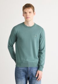 Green knit sweater with a round neckline, ribbed cuffs, and hem. Features a logo patch on the chest. Worn over blue jeans.