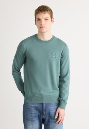 Groene gebreide trui met een ronde hals, geribbelde manchetten en onderkant. Bevat een logo-opdruk op de borst. Draagt over een blauwe spijkerbroek.