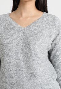Pull en maille gris avec décolleté en V, manches longues et texture douce. Le tissu présente un motif fin chiné.