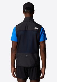 Black sleeveless vest with a lightweight design, featuring a mesh back panel, logo on the upper left, and reflective accents near the waist.