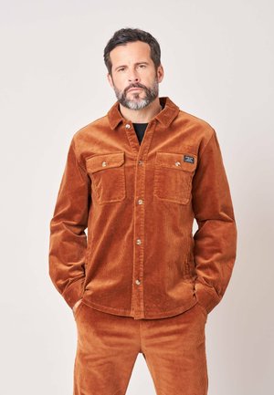 Man met kort donker haar en baard, gekleed in een bijpassend bruin corduroy overhemd en broek, staand met handen in de zakken tegen een lichte achtergrond.