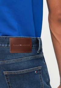 Κοντινό πλάνο πίσω τσέπης και ζωνάρι μπλε τζιν παντελονιών με καφέ δερμάτινο λογότυπο Tommy Hilfiger και κεντημένη λεπτομέρεια σημαίας.