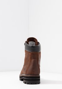 Botines de cuero marrón con cuello negro, superficie texturizada y suela contrastante con tacos en negro y marrón. La vista trasera muestra detalles de costura.