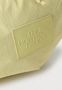 Κοντινή λήψη ενός ανοιχτό κίτρινου υφασμάτινου σακιδίου με ραμμένο ορθογώνιο έμπλαστρο με ανάγλυφο το λογότυπο και το έμβλημα της "THE NORTH FACE".