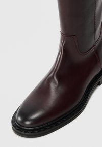 Bottine en cuir bordeaux avec une finition lisse, un bout rond et une semelle en caoutchouc noir texturé avec un détail de bord ondulé.