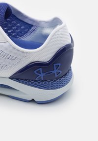 Scarpa sportiva bianca e blu navy con tomaia in mesh, collo imbottito, suola testurizzata e logo Under Armour blu prominente sul tallone. Marchio HOVR visibile.