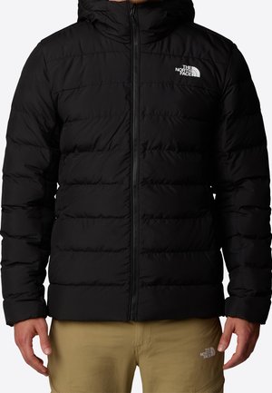 Homme portant une veste matelassée noire The North Face avec capuche et fermeture éclair devant, associée à un pantalon beige avec le logo The North Face.