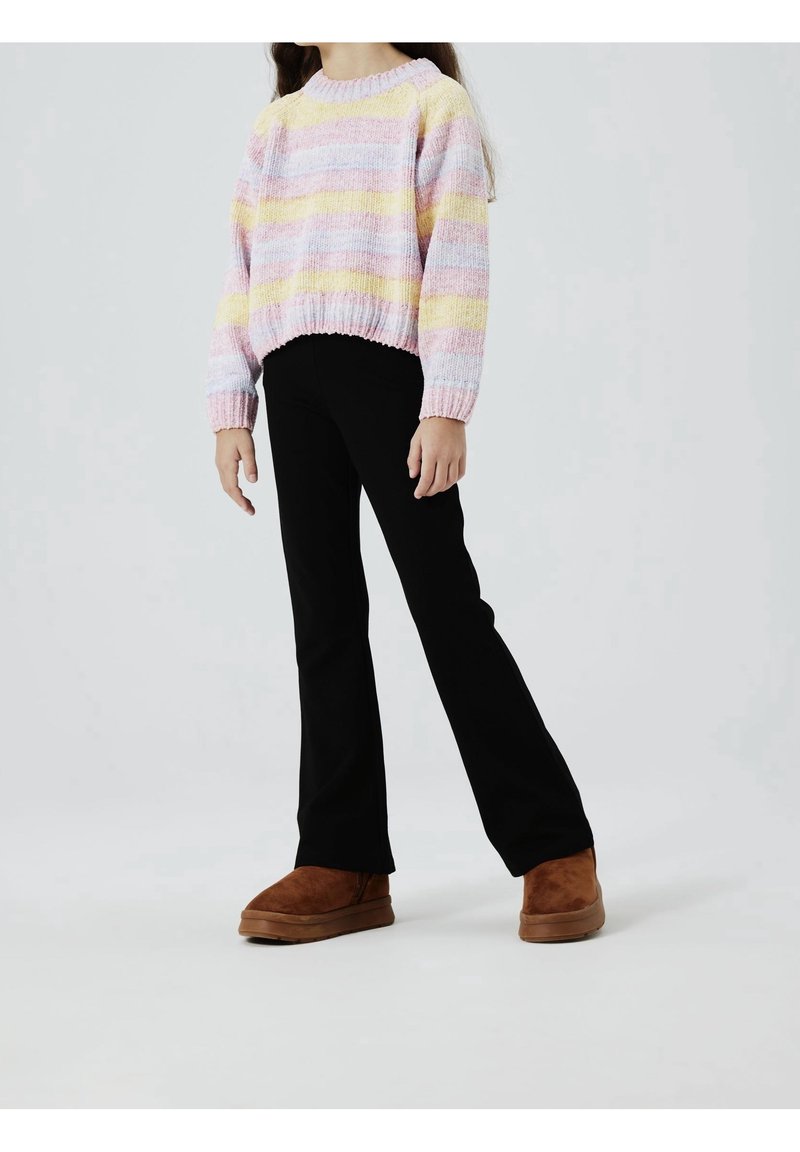 Enfant portant un pull rayé pastel, un pantalon noir et des chaussures en daim marron sans lacets, debout sur un fond uni.