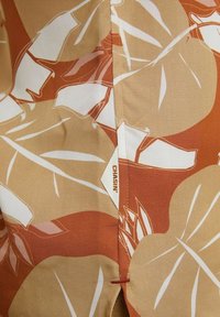 Camicia a maniche corte con un motivo di foglie nei toni dell'arancione e del beige, con cuciture tono su tono e un'etichetta con la scritta "CHASIN." sul lato.