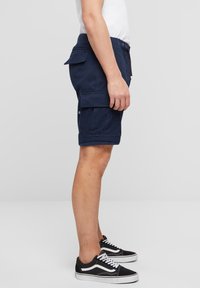 Pantaloni scurți cargo navy din țesătură durabilă, cu buzunare laterale, o croială lejeră și un tiv răsfrânt. Purtați cu teneși negri, accente albe.