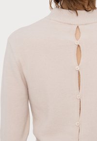 Vue arrière d'une personne portant un pull beige à col montant avec quatre boutons alignés verticalement ouvrant le dos.