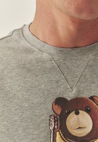 Szürke környakú pulóver, barna és arany maci grafikával, valamint a maci szőrzetén és gitárján található domború részletekkel.