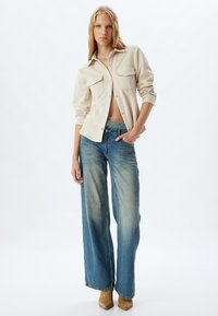 Camisa beige de botones con dos bolsillos en el pecho, combinada con vaqueros de pierna ancha de color azul claro con un diseño deslavado. Botas marrones hasta el tobillo completan el look.