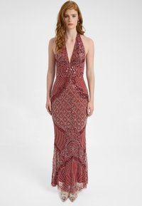 Abito lungo con collarino a halter in un profondo rosso, caratterizzato da intricate fantasie, una silhouette aderente e un orlo a sirena. Sandali con tacco trasparente completano l'outfit.