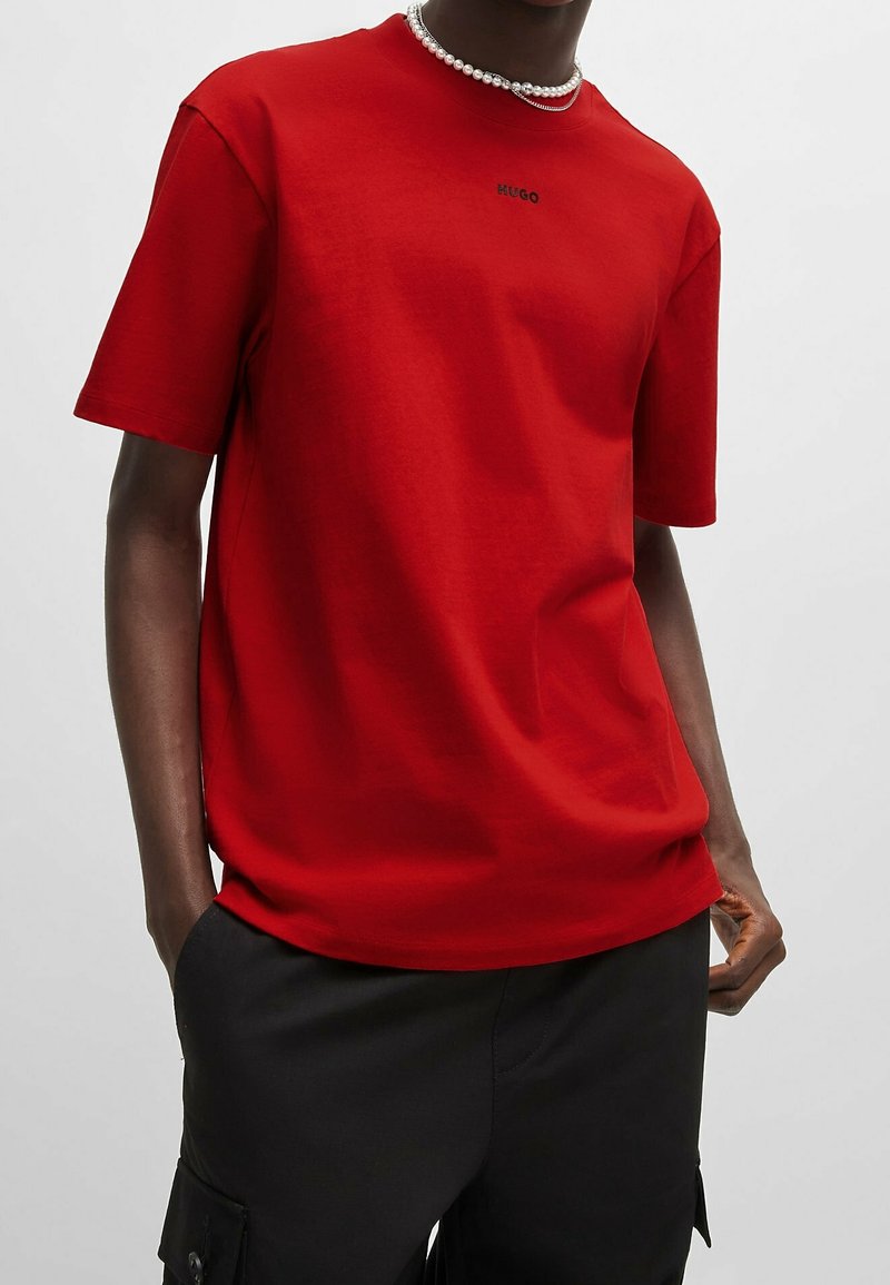 Personne portant une chemise rouge à manches courtes avec le texte "HUGO" et un collier de perles, associée à un pantalon noir avec des poches latérales.
